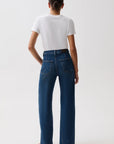 A model wearing a white short-sleeve crewneck t-shirt, high-waisted wide-leg blue jeans with a gold-tone belt, and black heeled sandals, standing against a plain light gray studio background, back