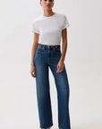 A model wearing a white short-sleeve crewneck t-shirt, high-waisted wide-leg blue jeans with a gold-tone belt, and black heeled sandals, standing against a plain light gray studio background.