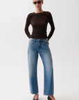 A woman modeling a dark brown long-sleeve knit top and wide-leg blue jeans with decorative silver studs down the sides, paired with black pointed-toe heels, against a plain white studio background.