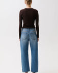 A woman viewed from behind wearing a dark brown long-sleeve top, blue wide-leg jeans, and black heels against a plain light gray studio background.