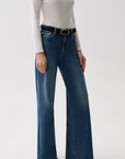 A model wearing a pair of dark blue wide-leg jeans with a black belt, paired with a white long-sleeve top and black pointed shoes, against a plain white studio background.