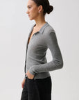A woman in profile wearing a fitted gray ribbed knit polo cardigan with a buttoned collar over a black top and black pants, photographed against a light gray studio background.