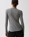 A woman in profile wearing a fitted gray ribbed knit polo cardigan with a buttoned collar over a black top and black pants, photographed against a light gray studio background, back