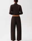 A model viewed from the back wearing a dark brown cropped long-sleeve shirt and matching wide-leg trousers, paired with black pointed-toe heels, against a plain light gray studio background.
