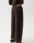 A person shown from the back wearing loose, wide-leg brown pants with a subtle small-scale check pattern, paired with black wedge sandals against a plain light gray studio background.