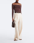 A model wearing a dark brown off-the-shoulder long-sleeve top, cream-colored pleated wide-leg trousers, a thin brown belt, and holding a small dark brown woven handbag against a plain white studio background.