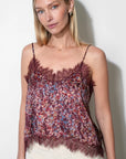 A woman wearing a burgundy lace camisole top with a red, blue, and white floral print, paired with cream-colored pants, standing against a plain white wall.
