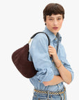 A model wearing a light blue denim shirt and jeans carries a dark brown suede crescent shoulder bag with gold-tone hardware against a plain white background.