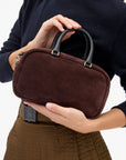 A person holding a small structured handbag in dark brown suede with black leather handles and gold-tone hardware, shown against a clean white background.