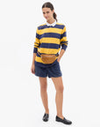 A woman modeling a yellow and navy blue horizontally striped long-sleeve rugby shirt with a white collar, paired with navy shorts, a brown belt bag, and black loafers, against a plain white background.
