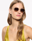 A woman with light brown hair wearing round gold-framed sunglasses with dark brown lenses and a shiny gold satin top, photographed against a plain white background.