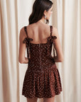 Woman wearing a brown polka dot dress with thin straps against a white curtain background, back view