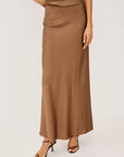 A woman wearing a long, flowing taupe-brown satin maxi skirt with a matching top and black strappy high-heeled sandals, photographed against a clean off-white studio background.