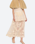 A model wearing a beige faux-fur capelet over a cream lace tiered midi skirt and nude high-heeled sandals, set against a plain light gray studio background.
