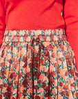 Close-up of a pleated skirt with a red, orange, and green floral print and a drawstring waist, paired with a bright red top.