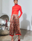 A model stands in a studio wearing a bright red mock-neck long-sleeve top, high-waisted wide-leg cropped pants with a red, orange, and green floral print, and red pointed-toe mule heels.
