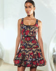 A woman modeling a sleeveless tiered mini dress with a dark navy background and a red, gold, and green floral print, standing in a brightly lit studio.