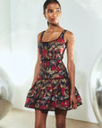 A model wearing a sleeveless tiered mini dress with a red, gold, and black floral jacquard pattern, standing in a brightly lit studio with white walls and reflective silver spheres.