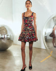 A model stands in a minimalist white studio space wearing a sleeveless mini dress with a red, black, and gold abstract pattern, paired with black pointed mule heels.