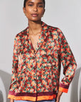 A woman wearing a long-sleeve satin-like button-front shirt jacket with an allover floral print in red, orange, green, and black, featuring contrasting orange and burgundy trim at the hem and cuffs, paired with light blue jeans.