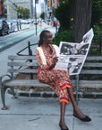 A woman sits on a city park bench reading a newspaper, wearing a colorful floral print dress with orange and yellow accents. The background shows a busy city street with cars, buildings, and trees.
