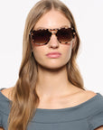 A woman with long light brown hair wearing oversized square tortoiseshell sunglasses with dark brown gradient lenses, set against a plain white background.