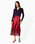A woman modeling a long-sleeve knit top with navy and red horizontal stripes paired with a red satin midi skirt featuring a lace hem, against a plain light gray background.