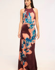 A woman modeling a sleeveless halter-neck maxi dress with a floral print in teal blue and peach-pink over a deep burgundy base, set against a plain white studio background.