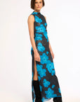 A model wearing a sleeveless, high-neck, floor-length dress with a black base and a vibrant blue floral pattern, styled with black knee-high boots against a plain white studio background.