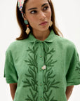 A woman wearing a green short-sleeve collared shirt with dark green embroidered leaf patterns down the front and a heart-shaped button, photographed against a plain light background.