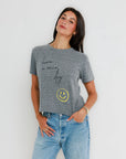 Woman wearing a gray t-shirt with a smiley face and text, standing against a white background