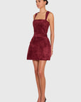 A woman modeling a sleeveless, square-neck mini dress in a deep burgundy velvet fabric, styled with black pointed-toe heels against a solid light gray background.