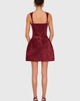 A woman viewed from the back wearing a sleeveless burgundy mini dress with shoulder straps and a fitted waist, paired with black high-heeled sandals, against a light gray studio background.