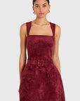 A model wearing a sleeveless, square-neck mini dress in a deep burgundy velvet-like fabric, featuring a matching belt with a square buckle and front patch pockets, set against a plain light gray background.