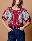 A woman modeling a short-sleeved blouse with a red and white floral pattern, paired with dark blue jeans and a black belt, set against a solid tan background.
