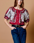 A woman modeling a short-sleeve blouse with a red and white floral pattern, paired with dark blue jeans and a black belt, against a solid tan background.