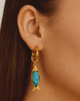 Gold earring with turquoise stone fish charm worn by a person