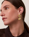 A close-up side profile of a woman wearing a gold spiral drop earring, showing the earring against her ear and neck.