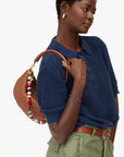 Woman holding a woven handbag, wearing a blue top and green pants on a white background
