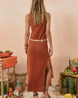 Woman in a rust-colored dress standing in a tropical setting with fruits and decor. back view