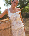 Woman in a white lace tank and pants holding a woven basket outdoors.