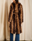 A woman stands with her back to the camera wearing a long brown faux fur coat with a white spotted pattern, blue jeans, and black ankle boots, set against a cream curtain backdrop.