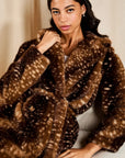 A woman wearing a plush brown faux fur coat with a white spotted pattern, posed against a neutral beige background.