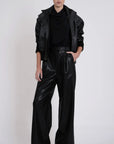 A model stands against a light gray studio background wearing a black faux-leather jacket and matching high-waisted, wide-leg pants with a glossy finish.