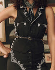 A close-up of a model wearing a black sleeveless vest and matching skirt set with white embroidered details, styled with a black and white patterned neck scarf and a black belt.