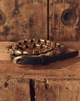 A black leather belt with a large gold buckle and a matching gold chain bracelet, styled on a rustic wooden surface with a matching wooden background.