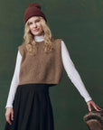 Woman wearing a brown knitted vest, white shirt, and dark skirt against a green background