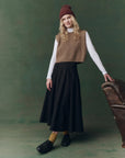 Woman wearing a brown sweater vest, black skirt, and maroon beanie against a green background, full body