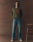 The college sweatshirt rodeo star army green on a model wearing jeans with a brown background, front view