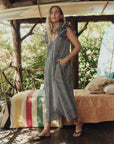 Woman in a blue checkered dress standing in a rustic outdoor setting with greenery. side view